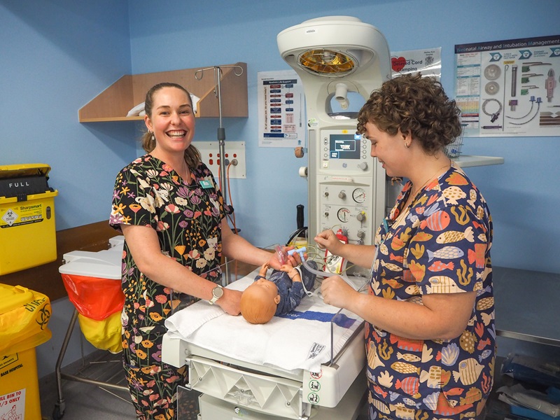 A midwife in scrubs stands over a baby cot with a trainee