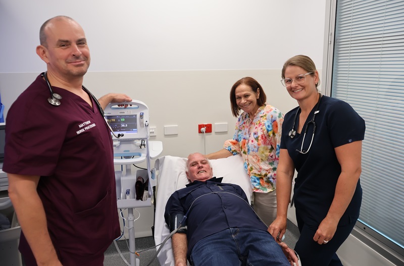 Two nurses stand with a cardiac patient laying on a clinic bed with his wife bedside him