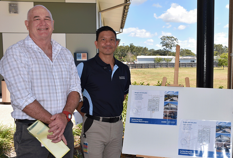 Two men stand in front of blueprints for a hospital