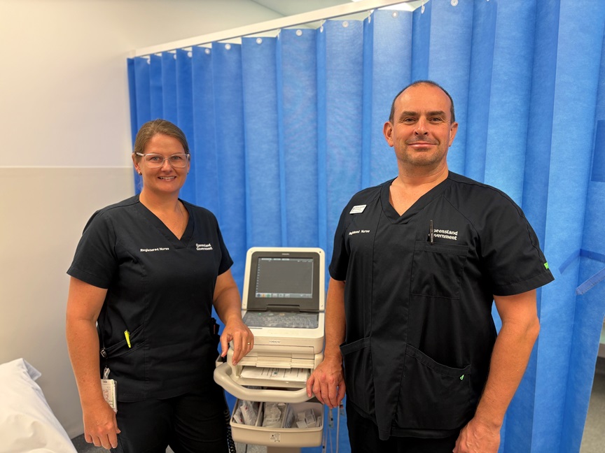 A male and female nurse stand beside a blood pressure machine