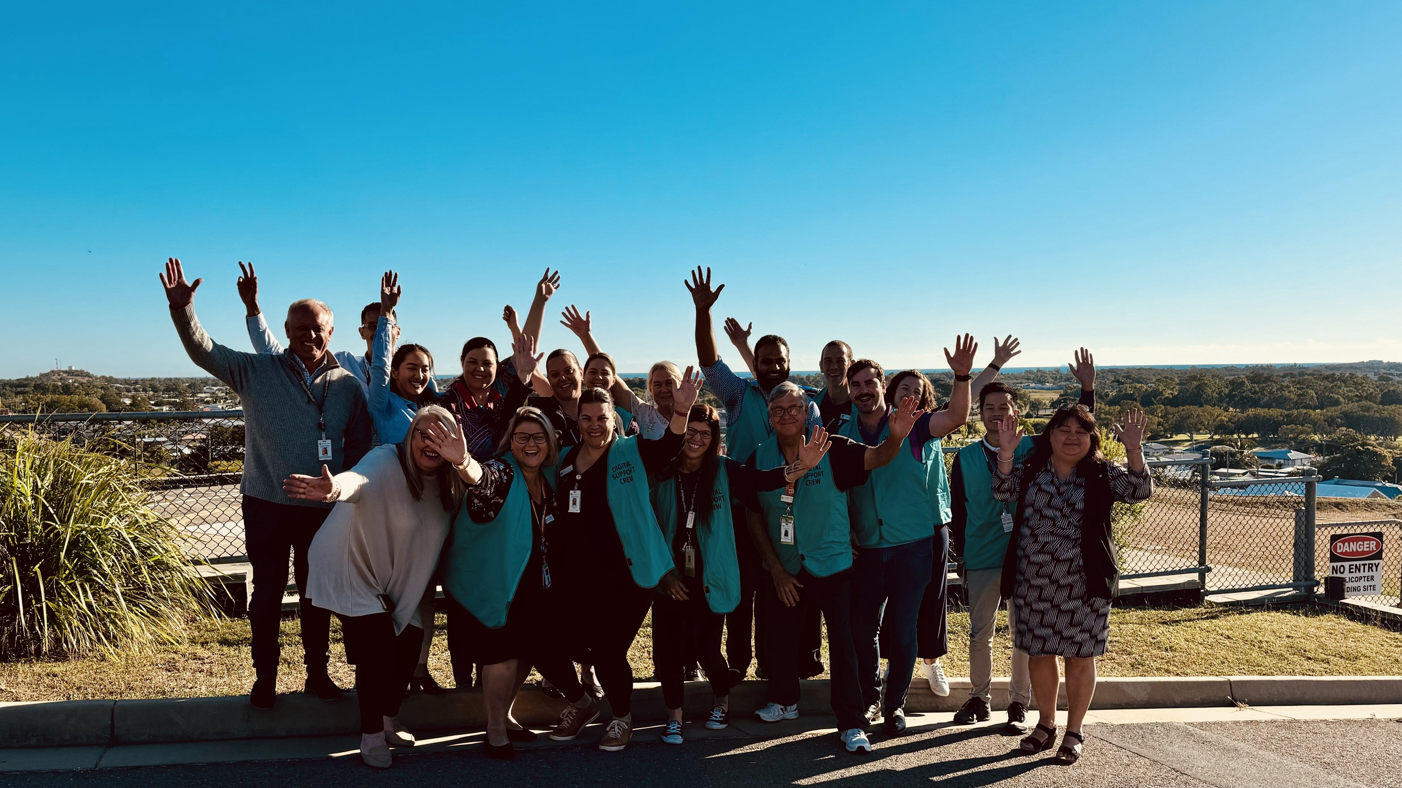 Group of Bowen Hospital staff celebrate by throwing their hands in the air outside with view behind them.